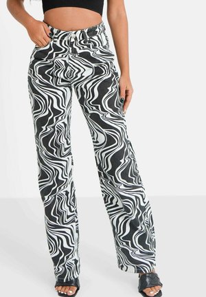 Vrouw draagt zwarte en witte broek met hoog taille en een marmerachtig patroon, een zwart crop top en zwarte open-toe hakken, staand met één hand in de zak.