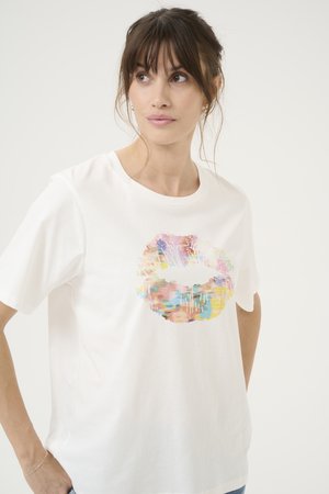 Kvinde med mørkt hår iført en hvid t-shirt med et stort farverigt abstrakt læbemotiv, stående med den ene hånd på hoften.