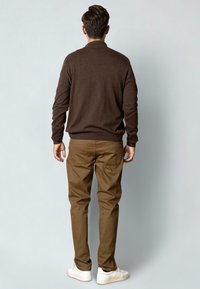 Mies, joka katsoo pois, päällään ruskea neulepaita, khaki-housut ja valkoiset lenkkarit, yksinkertaista vaaleanharmaata taustaa vasten.