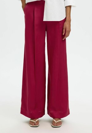 Femme portant un pantalon bordeaux à jambes larges et des sandales blanches à bout carré, avec une chemise blanche à manches longues partiellement visible.