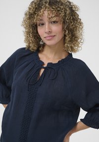 Navy blue blouse in lightweight fabric with a round neckline, tie detail, and textured trim down the center. Puff sleeves with elastic cuffs.