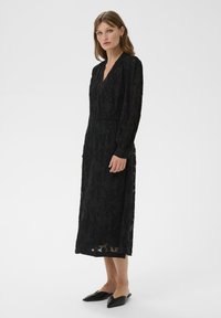 Schwarzes Spitzenkleid mit gemusterter Textur, V-Ausschnitt, langen Ärmeln und knielangem Saum. Getragen mit spitzen schwarzen Ballerinas.