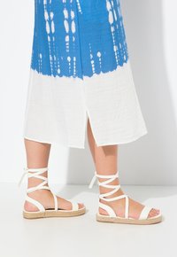 Vita remsandaler med jutesula, ankelsnören och minimalistisk design, kombinerade med en klänning i blå och vit mönstring.