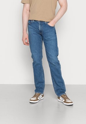 Muž v modrých džínech s rovnými nohavicemi, béžovém tričku a béžovo-bílých teniskách, stojící s jednou rukou v kapse.