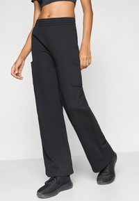 Μαύρο φαρδύ παντελόνι φόρμας με ελαστική μέση, δύο πλαϊνές τσέπες cargo και λεία υφή, συνδυασμένο με μαύρα αθλητικά παπούτσια.