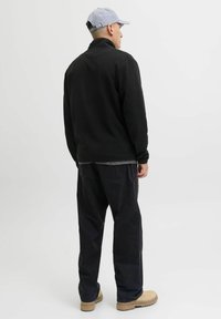 Giacca in pile nera con colletto alto, indossata sopra una camicia grigia, abbinata a pantaloni scuri e stivaletti beige chiaro. La vista posteriore mostra un design minimale.