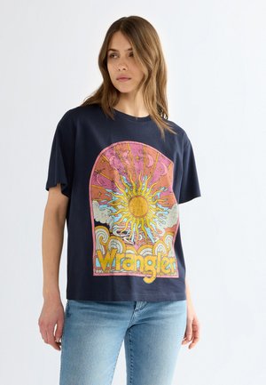Femme portant un t-shirt bleu marine avec un motif coloré de soleil, phases de la lune, nuages et texte "Wrangler", associé à un jean bleu clair.