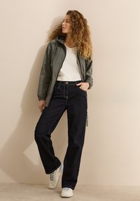 Chaqueta: gris, ligera, con cremallera frontal y capucha; camiseta blanca; pantalones de mezclilla oscuros con detalles de costura; zapatillas blancas con acentos en azul claro.