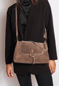 Taupe suede schoudertas met een gestructureerd ontwerp. Heeft een gouden ronde accenten en een gevlochten detail op de flap. Inclusief verstelbare riem.