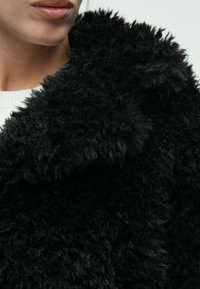 Μαύρο παλτό από υλικό fleece με υφή, υψηλός γιακάς και μαλακό, απαλό ύφασμα. Κοντινή εικόνα αποκαλύπτει κυματιστές ίνες και παχύ υλικό.