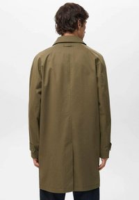 Olivgrön trenchcoat med ryggslits, axelvaddar och knappade manschetter. Slät tygtextur med avslappnad passform.