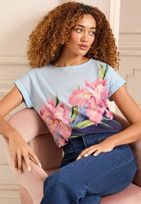 Šviesiai mėlynas trumpomis rankovėmis viršus, puošiamas dideliais rožiniais gėlių raštais, suapvalintu iškirpimu ir laisvu siluetu, derinamas su tamsiomis džinsinėmis kelnėmis.