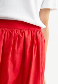 Gonna rossa leggera con vita arricciata. Realizzata in tessuto morbido, presenta pieghe verticali per una consistenza fluida. Indossata con una maglietta bianca.