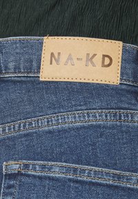 Pantalones vaqueros de mezclilla con un parche de cuero marrón claro etiquetado "NA-KD" en la cinturilla; tela azul, costuras visibles y detalle del quinto bolsillo.