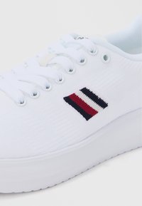 Bela platnena superga s ploskim podplatom, okrolim prstom in črtastim logotipom v mornarsko modri, rdeči in beli barvi na strani. Zapenjanje z vezalkami s ploskim vrvico.