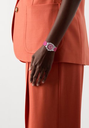 Rosa Uhr mit einem strukturierten Armband, runden silbernen Gehäuse und detailliertem rosa Zifferblatt. Wird an einer Hand getragen, kombiniert mit einem orangefarbenen Sakko.