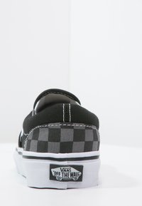 Scarpa slip-on a scacchi neri e grigi con suola bianca e cuciture bianche. Presenta il logo Vans sul tallone. Materiale in tessuto con finitura testurizzata.