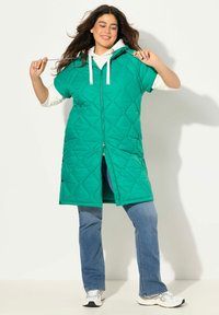 Grön vadderad väst med dragkedja och huva, med kortärmad design. Bärs över en vit hoodie och blå jeans, med sneakers.