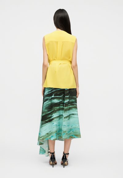 Femme aux longs cheveux foncés portant un haut jaune sans manches et une jupe asymétrique à motifs vert-bleu, debout de dos.