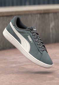 Šedé semišové sneakersy Puma s bílým logem a podešví, zavázané, zavěšené nad kamenným povrchem s rozmazaným pozadím.