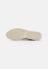 Sneaker outsole with a textured rubber surface featuring a zigzag tread pattern, cream color with a logo in the center.