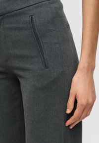 Κοντινό πλάνο ενός χεριού που ακουμπά δίπλα σε σκούρο γκρι παντελόνι ραμμένο εμπεριστατωμένα με κάθετη τσέπη στην πλευρά.