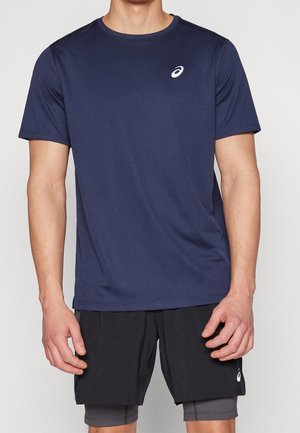 Man draagt een donkerblauw sportshirt met korte mouwen en zwarte gelaagde shorts, staand tegen een effen lichte achtergrond.