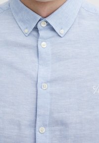 Mies, joka käyttää vaaleansinistä nappikauluspaitaa, jossa on napitettu kaulus ja valkoiset napit, näyttäen paidan etuosaa ja yläkaulaa.