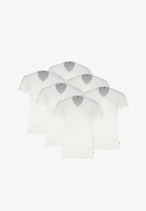 Mann trägt ein schlichtes weißes V-Neck-T-Shirt und graue Boxershorts, steht vor einem einfarbigen hellen Hintergrund.