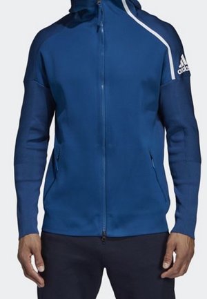 Blauer Adidas Zip-Up-Hoodie mit weißem Streifen auf der Schulter und Logo am oberen linken Arm, getragen von einer Person mit entspannt an den Seiten liegenden Händen.