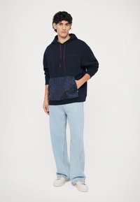 Marineblå hettegenser med røde snorer og mønstret lomme; kombinert med lyseblå jeans og hvite joggesko. Laget av bomull, avslappet passform.