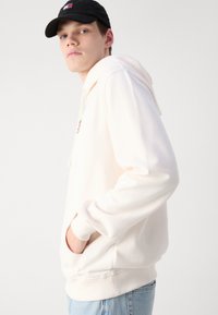 Light pink hoodie made of soft fabric, featuring a front pocket, drawstring hood, and a small logo on the chest. Worn with blue jeans.