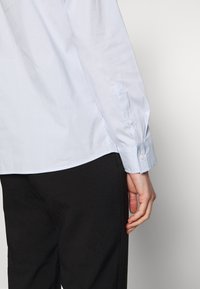 Camicia a righe azzurro chiaro con maniche lunghe, polsini abbottonati e orlo curvo, abbinata a pantaloni neri su misura. Tessuto in cotone morbido.