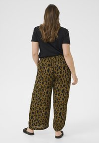Mujer con cabello largo vistiendo una camiseta negra, pantalones sueltos de estampado de leopardo y sandalias negras, de pie de espaldas sobre un fondo blanco.