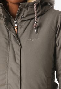 Zbliżenie na osobę noszącą zieloną zimową kurtkę z kapturem, z zamkiem błyskawicznym, guzikami i sznurkiem ze metalowymi końcówkami.