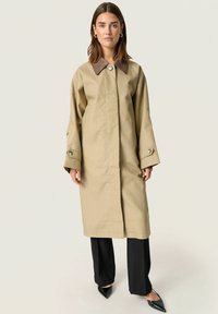 Beige színű trench coat barna gallérral, elöl gombos zárással és széles ujjakkal. Fekete nadrág és hegyes fekete cipő fölött viselve.