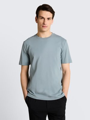 Ung mand med kort mørkt hår iført en lyseblå kortærmet t-shirt og sorte bukser, stående med hænderne i lommerne mod en ensfarvet baggrund.