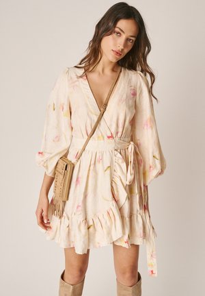 Vrouw gekleed in een lichte bloemenprint wikkeljurk met ballonmouwen, een beige crossbodytas en suède laarzen tot aan de knie tegen een effen achtergrond.