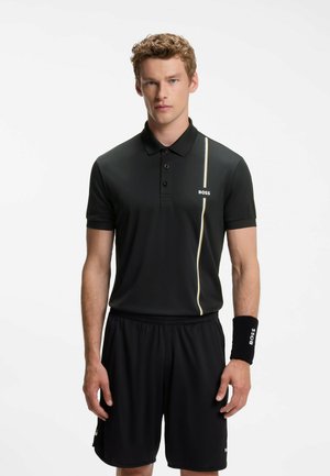 Giovane uomo che indossa una polo nera BOSS e pantaloncini con braccialetto nero, in piedi davanti a uno sfondo chiaro e uniforme.