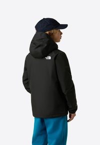 Crna vodootporna jakna s kapuljačom, s bijelim logom na leđima. Kombinirana s plavim hlačama i tamnoplavom kapom.