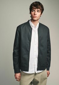 Fekete bomber dzseki cipzárral az elején és bordás mandzsettával, egy fehér gombos ing fölött viselve. Sima textúra, minimalista design.