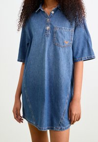 Robe-chemise en jean bleu moyen avec manches courtes, col, patte de boutonnage à trois boutons et une poche poitrine ornée d'un détail logo en forme de cœur cousu.