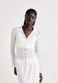 Black woman with braided hair wearing a white textured buttoned shirt and white eyelet skirt against a plain light background.