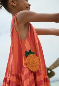 Abito a strisce rosse e arancioni con un accessorio arancione arrotondato e texturizzato, con foglie verdi attaccate da una tracolla intrecciata.