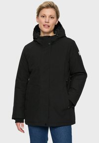 Vrouw in een zwarte gewatteerde winterjas met capuchon en een paarse jeans, staand tegen een effen lichte achtergrond.