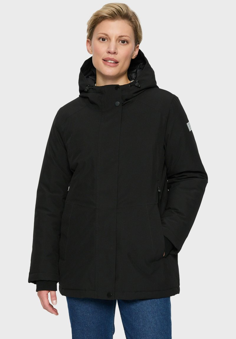 Vrouw in een zwarte gewatteerde winterjas met capuchon en een paarse jeans, staand tegen een effen lichte achtergrond.