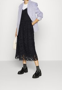 Robe midi en dentelle noire avec motif floral, superposée à un T-shirt blanc, accompagnée d'un blazer bleu clair et de chaussures à plateforme noires.