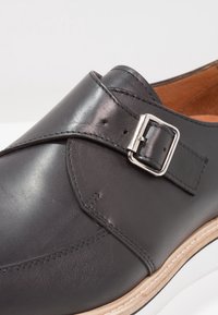 Scarpa in pelle nera con finitura liscia, dotata di una fibbia argentata e cuciture in contrasto. Suola chiara con una leggera curvatura.