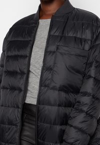 Chaqueta acolchada negra con diseño acolchado, cierre con cremallera, un cuello de canalé y un bolsillo frontal. Textura ligera, suave y cosida.