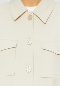 Veste boutonnée beige clair avec un tissu texturé, deux poches poitrine et des boutons ronds avec un éclat subtil. Design de col classique.